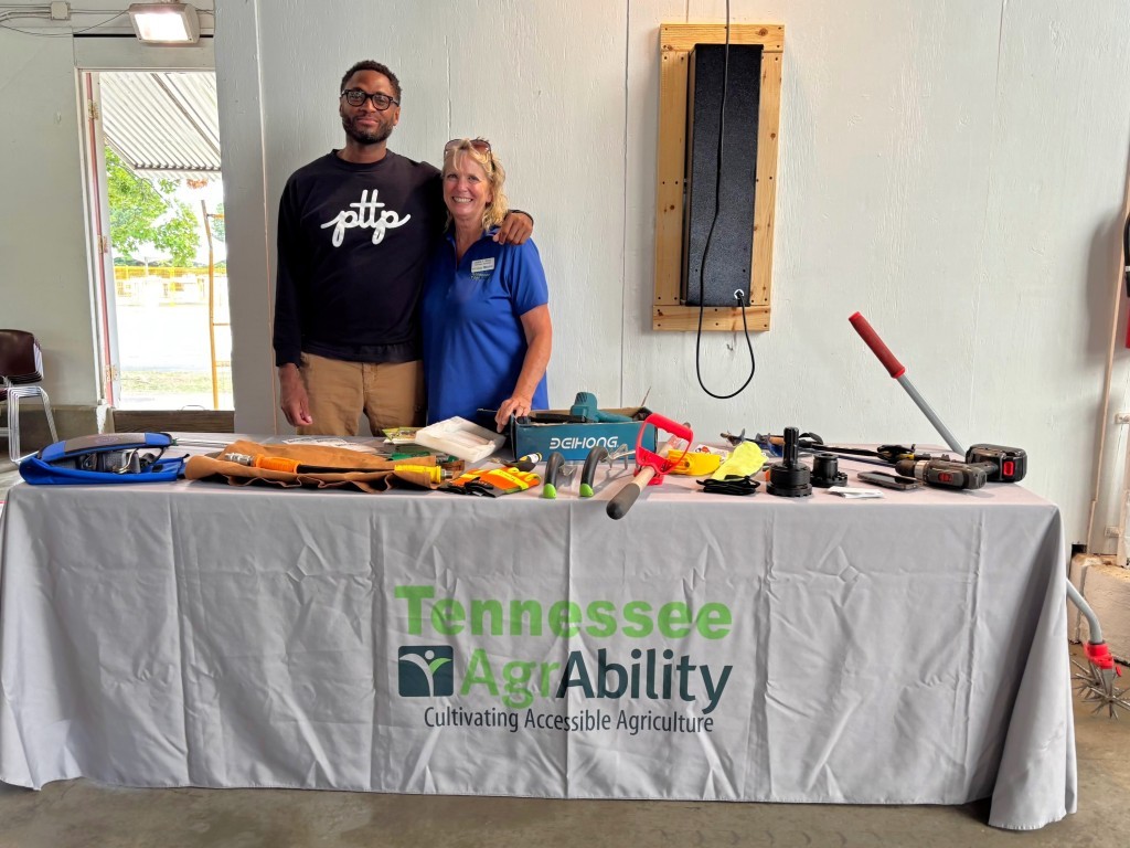 A man and a woman standing behind a TN AgrAbility display table with small AT items on it